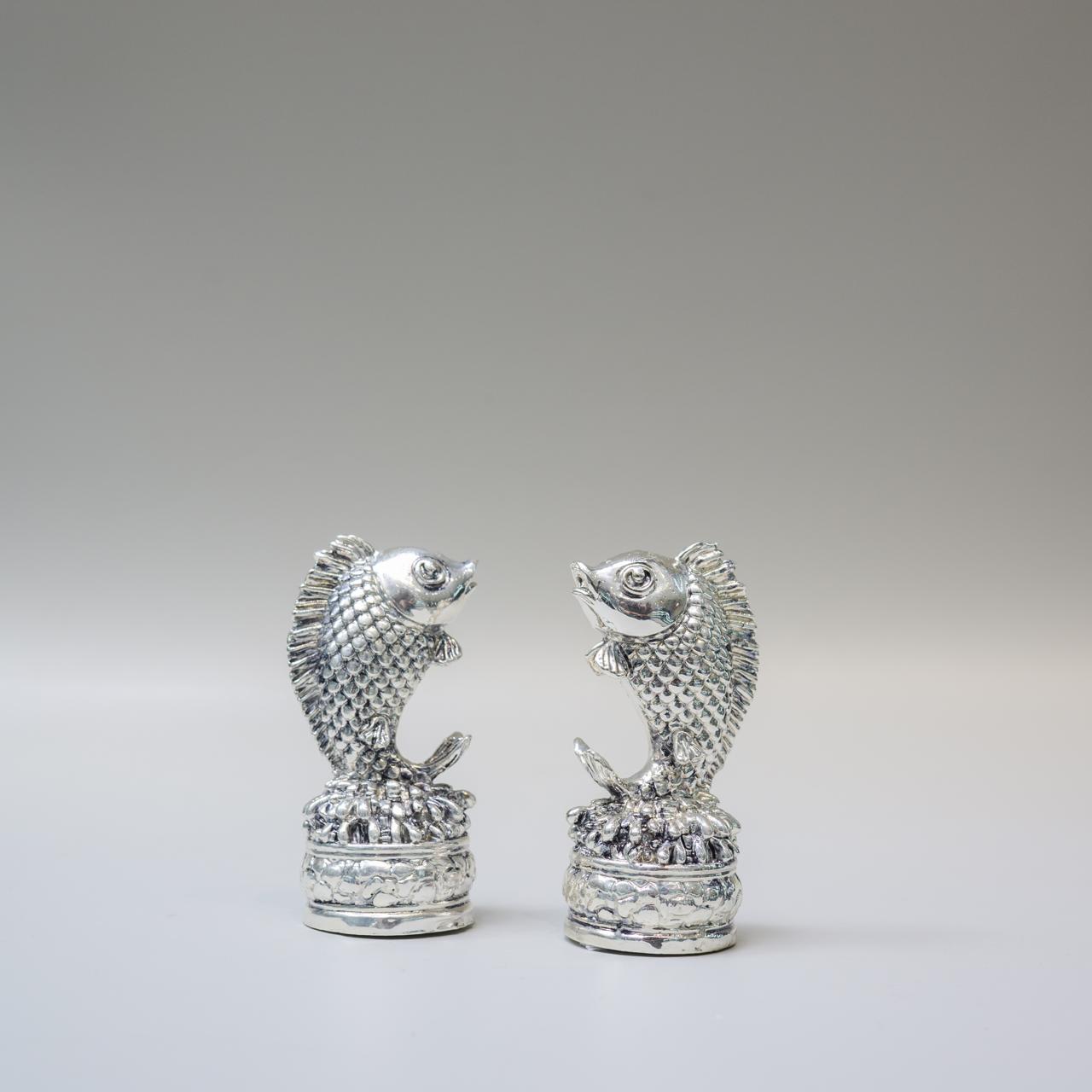 A pair of small silver fish figurines standing upright on textured cylindrical bases, photographed against a smooth light gray studio background.