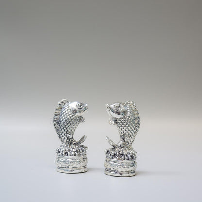 A pair of small silver fish figurines standing upright on textured cylindrical bases, photographed against a smooth light gray studio background.