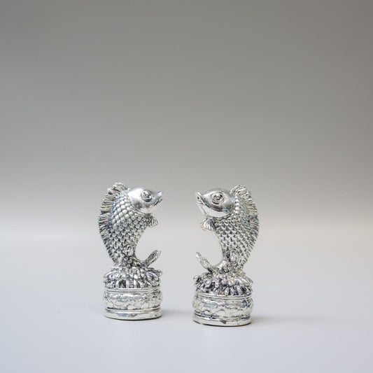 A pair of small silver fish figurines standing upright on textured cylindrical bases, photographed against a smooth light gray studio background.